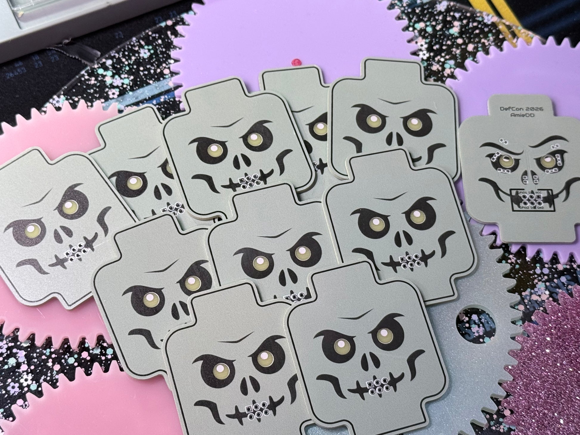 Several unassembled skeleton minifig-inspired DefCon SAO PCBs arranged on a pink and purple background, showing the skull-face artwork and one reverse-side board.